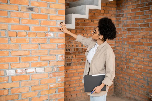 Afrikanisch-amerikanischer Immobilienmakler mit Folder, der Ziegelwand in Haus mit unfertigem Interieur berührt — Stockfoto