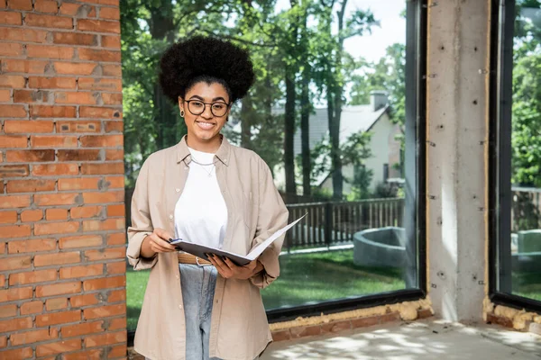 Joyeux agent immobilier afro-américain avec dossier regardant la caméra dans la maison avec de grandes fenêtres — Photo de stock