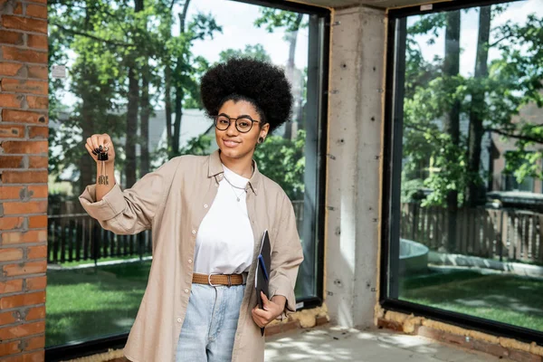 Agente inmobiliario afroamericano positivo que sostiene la carpeta y las llaves en la casa de campo moderna de la ciudad - foto de stock