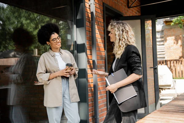 Courtier immobilier blonde parler à une femme afro-américaine avec du café pour aller près de chalet — Photo de stock