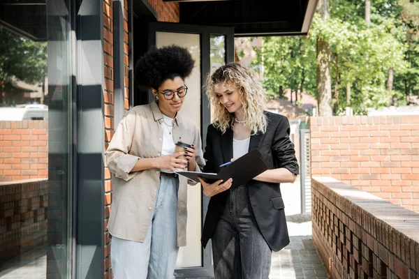Real estate broker showing documents to african american woman with takeaway drink near house — Stock Photo