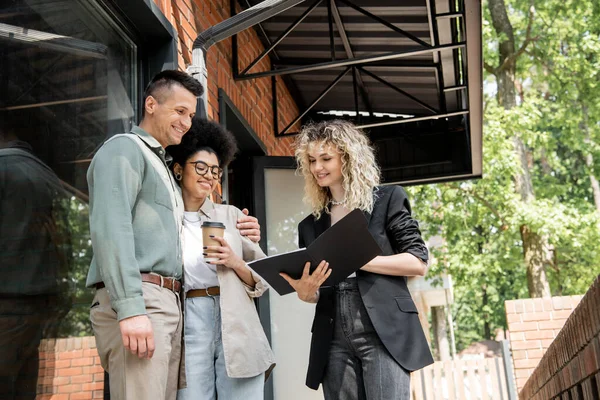 Smiling real estate agent showing documents to interracial couple with paper cup near cottage — Stock Photo