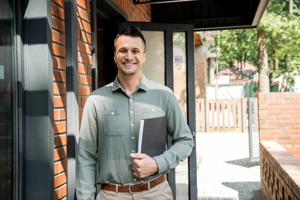 Successful and happy real estate agent with folder looking at camera near modern cottage — Stock Photo