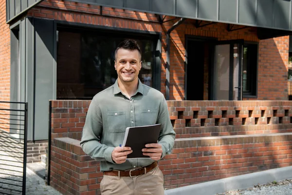 Confident real estate agent holding folder and looking at camera near modern house on street — Stock Photo