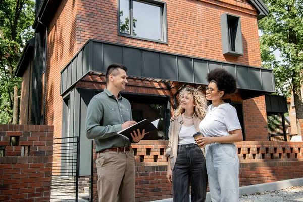 Positive real estate agent talking to interracial lesbian couple with coffee to go near city cottage — Stock Photo