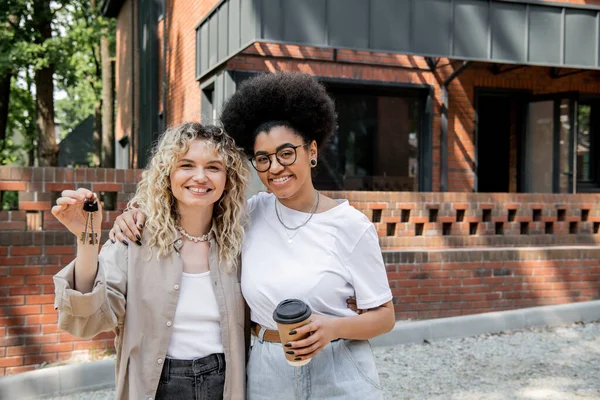 Cheerful interracial lesbian couple with keys and coffee to go looking at camera near private house — Stock Photo