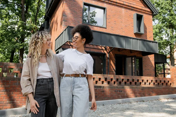 Joyful multiethnic lesbian couple looking at each other next to private modern cottage — Stock Photo