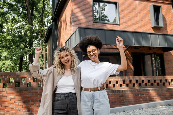 Emocionadas mujeres lesbianas multiculturales regocijándose y mostrando gesto de victoria cerca de casa privada - foto de stock