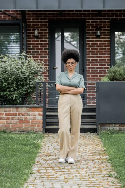 Piena lunghezza di sorridente agente immobiliare africano americano in piedi con le braccia piegate vicino cottage — Foto stock