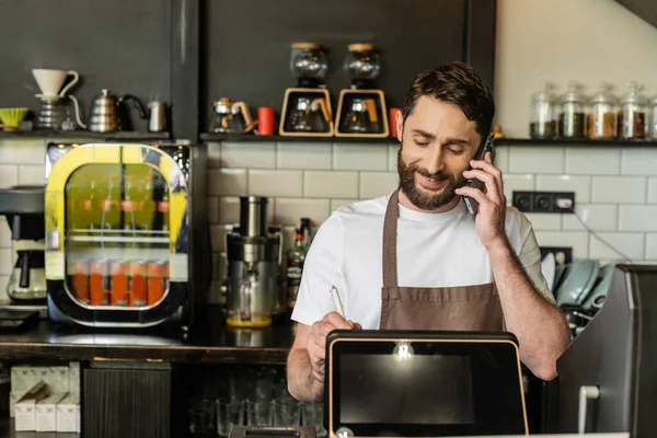 Sorridente barista no avental falando no smartphone e usando tablet digital no café — Fotografia de Stock