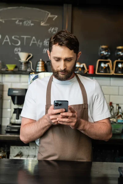 Bearded barista in apron using smartphone while standing near bar and working in coffee shop — Stock Photo