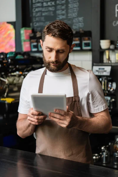 Barista barbudo en delantal usando tableta digital mientras trabaja y está de pie cerca del bar en la cafetería - foto de stock