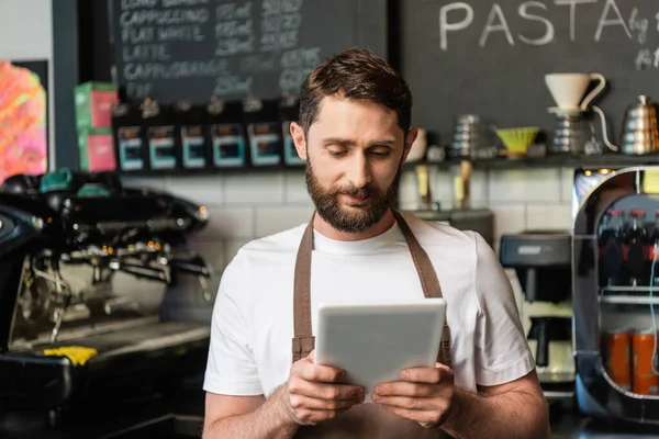 Бариста в фартуке с цифровым планшетом во время работы и стояния возле бара в кофейне — стоковое фото