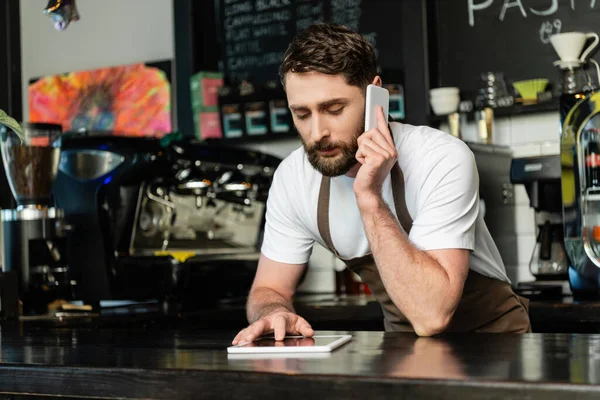 Barista dans tablier parler sur smartphone et en utilisant tablette numérique sur bar dans un café — Photo de stock