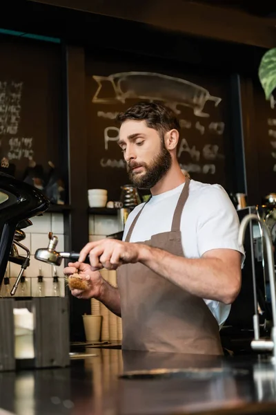 Bearded barista in apron cleaning coffee machine holder with brush in coffee shop — Stock Photo