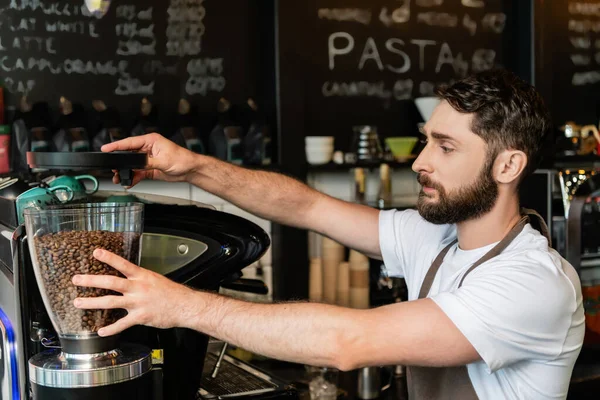 Barista barbudo em avental usando moedor de café perto da máquina enquanto trabalhava em café — Fotografia de Stock