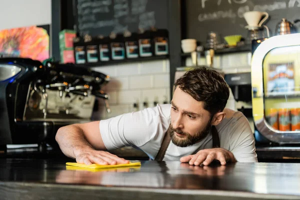 Barista barbudo enfocado en la barra de limpieza del delantal y sosteniendo el trapo mientras trabaja en la cafetería - foto de stock