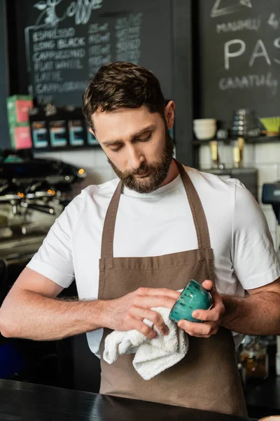 Barista barbudo y enfocado en delantal taza de limpieza con toalla mientras trabaja en la cafetería - foto de stock