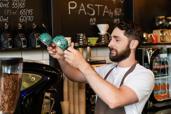 Barista barbu souriant dans tablier tenant tasses près de la machine à café tout en travaillant dans un café — Photo de stock