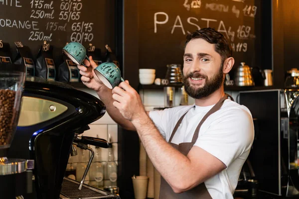 Positiver bärtiger Barista in Schürze, der in die Kamera blickt und Tassen in der Nähe der Kaffeemaschine im Café hält — Stockfoto