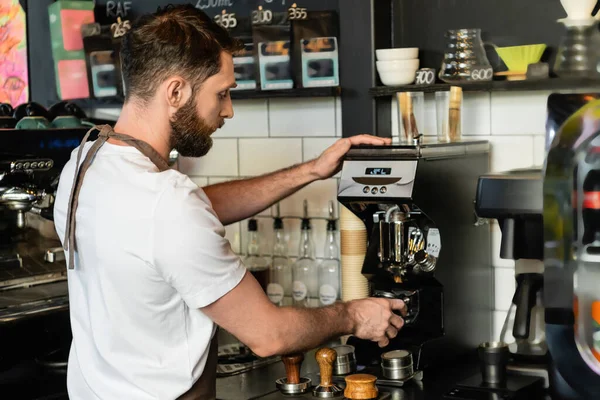 Vista lateral do barista no avental derramando café no suporte perto da máquina de café enquanto trabalhava no café — Fotografia de Stock
