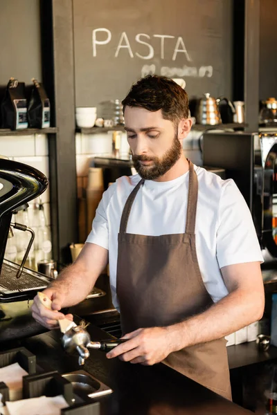 Bearded barista in apron cleaning holder with brush near coffee machine while working in cafe — Stock Photo