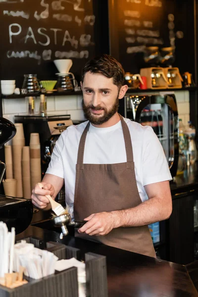 Smiling barista in apron looking at camera while cleaning coffee machine holder with brush in cafe — Stock Photo
