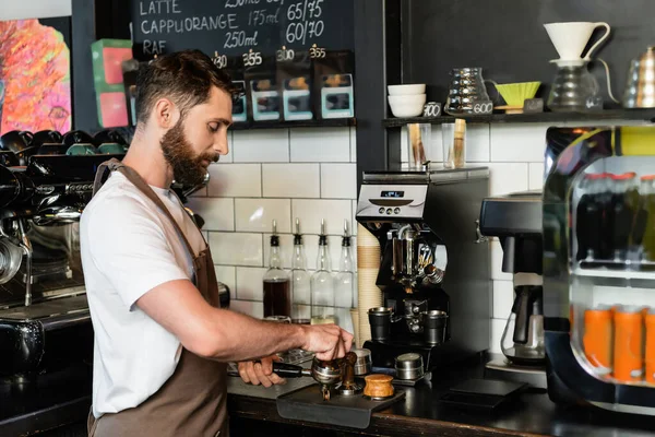 Vista lateral do barista barbudo no avental pressionando café no suporte enquanto trabalhava no café — Fotografia de Stock