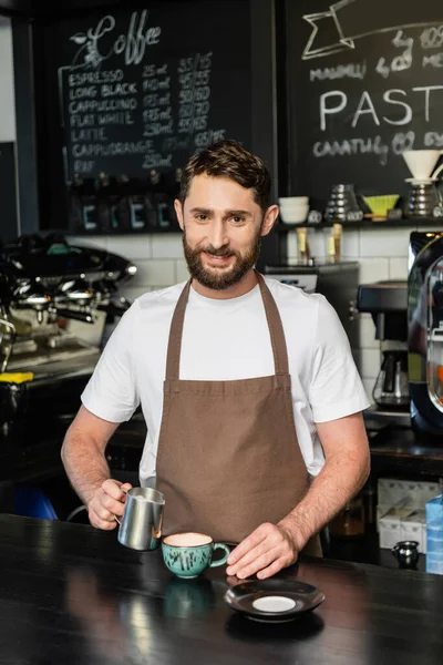 Sorridente barista no avental segurando jarro com leite perto cappuccino na xícara no café — Fotografia de Stock