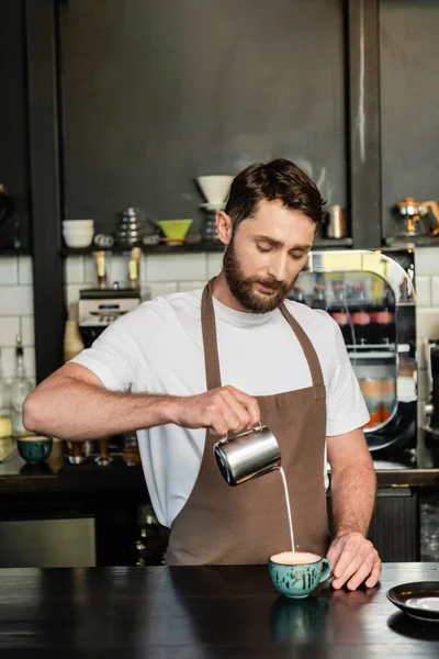 Bärtiger Barista in Schürze, der Milch aus dem Krug gießt, während er Cappuccino auf der Theke im Café zubereitet — Stockfoto