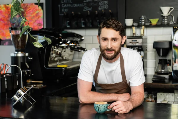 Barista barbu souriant dans le tablier regardant la caméra près de la tasse de cappuccino sur le bar dans le café — Photo de stock