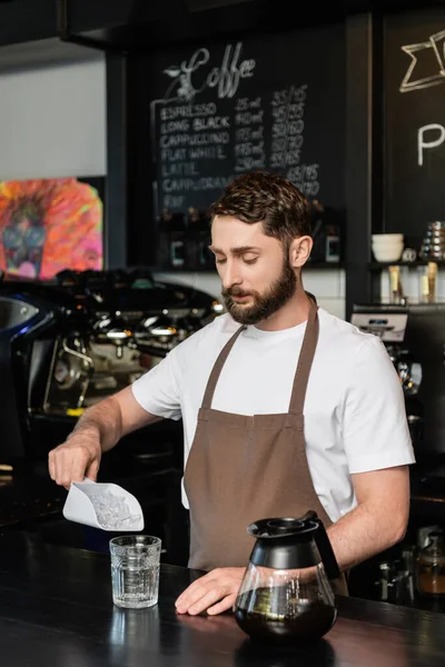 Barista in apron pouring ice cubes in glass near coffee pot on bar in coffee shop — Stock Photo