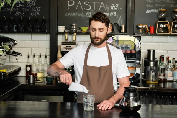 Barista gießt Eiswürfel in Glas neben Kaffeekanne auf Bar, während er im Coffeeshop arbeitet — Stockfoto