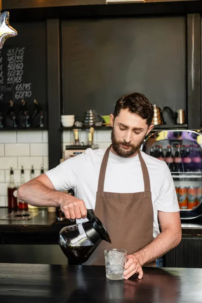 Barista en delantal verter el café de la olla en el vaso con cubitos de hielo en el bar en la cafetería - foto de stock