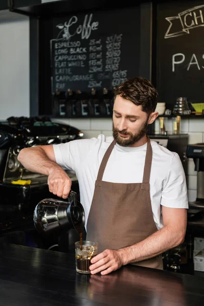 Bärtiger Barista, der Kaffee aus Kanne in Glas mit Eiswürfeln gießt, Bar im Café, Eiskaffee — Stockfoto