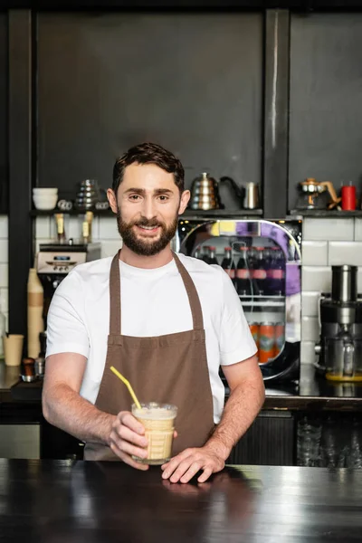 Улыбающийся бариста в фартуке держит холодный кофе напиток с соломой и смотрит в камеру в кафе — стоковое фото