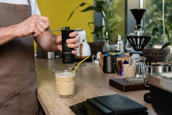 Vista cortada de barista em avental moagem de café perto de bebida de café frio em vidro no café — Fotografia de Stock