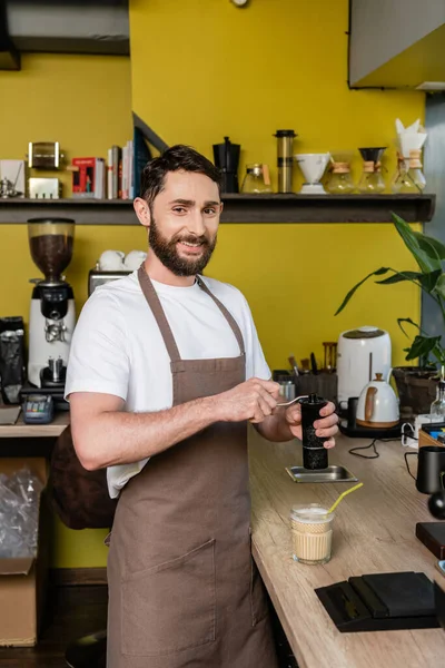 Cheerful barista in apron grinding coffee and looking at camera near cold coffee drink in cafe — Stock Photo