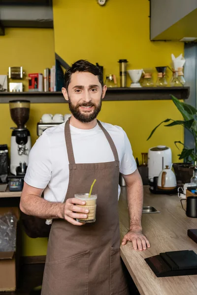 Barista positivo em avental segurando bebida de café frio em vidro e sorrindo para a câmera no café — Fotografia de Stock