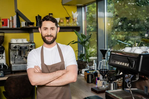Barista sonriente en delantal cruzando brazos y mirando a la cámara cerca de la máquina de café en la cafetería - foto de stock