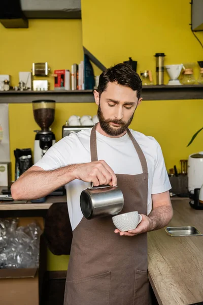 Сфокусований бариста в фартусі, що заливає молоко в чашці з кавою під час роботи в кав'ярні — стокове фото