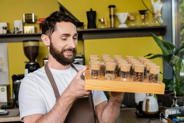Улыбающийся бородатый бариста в фартуке держит кофейные зерна в банках во время работы в кафе — стоковое фото