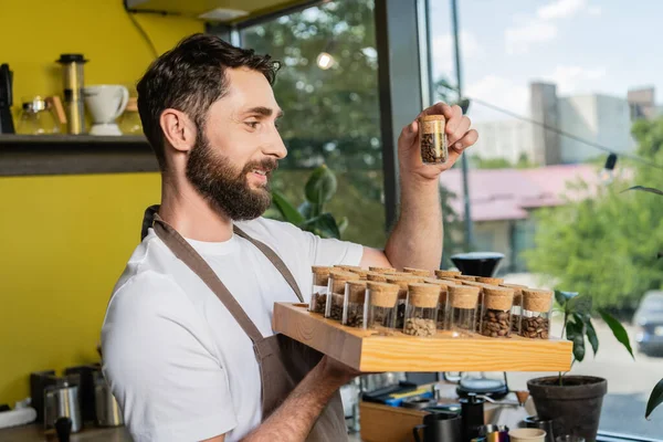 Barista barbu positif tenant des pots avec des grains de café tout en travaillant dans un café — Photo de stock