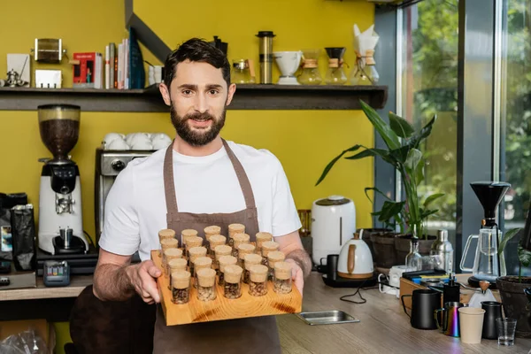 Smiling barista in apron holding jars with coffee beans and looking at camera in coffee shop — Stock Photo