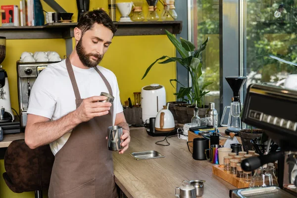Barista barbudo em avental segurando jarros de metal ao fazer café no café — Fotografia de Stock