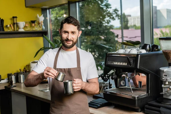 Cheerful bearded barista in apron holding pitchers near coffee machine in coffee shop — Stock Photo
