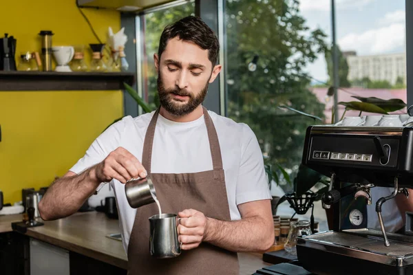 Barista barbudo enfocado en delantal sosteniendo jarras cerca de la máquina de café en la cafetería - foto de stock