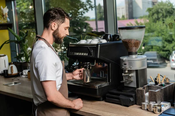 Vue latérale du lait barbu moussant barista tout en travaillant avec une machine à café dans un café — Photo de stock