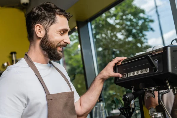Вид сбоку улыбающегося и бородатого бариста в фартуке с использованием кофемашины во время работы в кафе — стоковое фото
