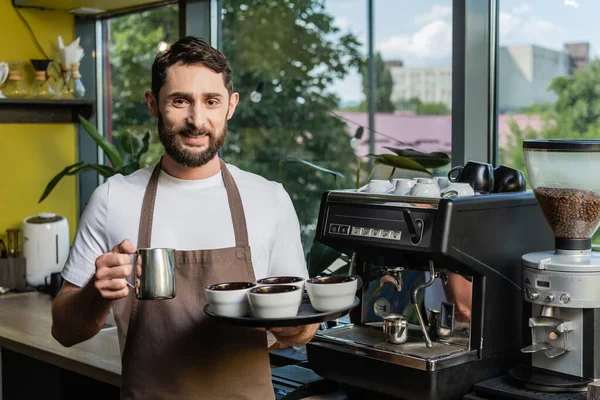 Barista sonriente en delantal sosteniendo jarra y tazas cerca de la máquina de café en la cafetería - foto de stock